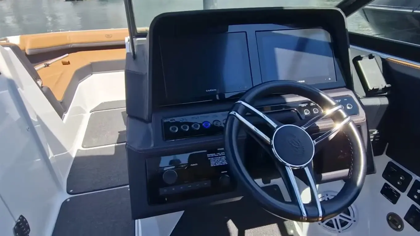 Boat cockpit with a steering wheel, dual Garmin displays, and control buttons, set against a water backdrop.